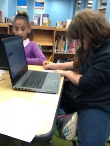 One girl looking at a computer, another girl writing down an answer on paper