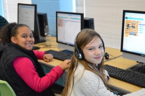 Two girls sitting at computers working on coding, turning to smile at camera